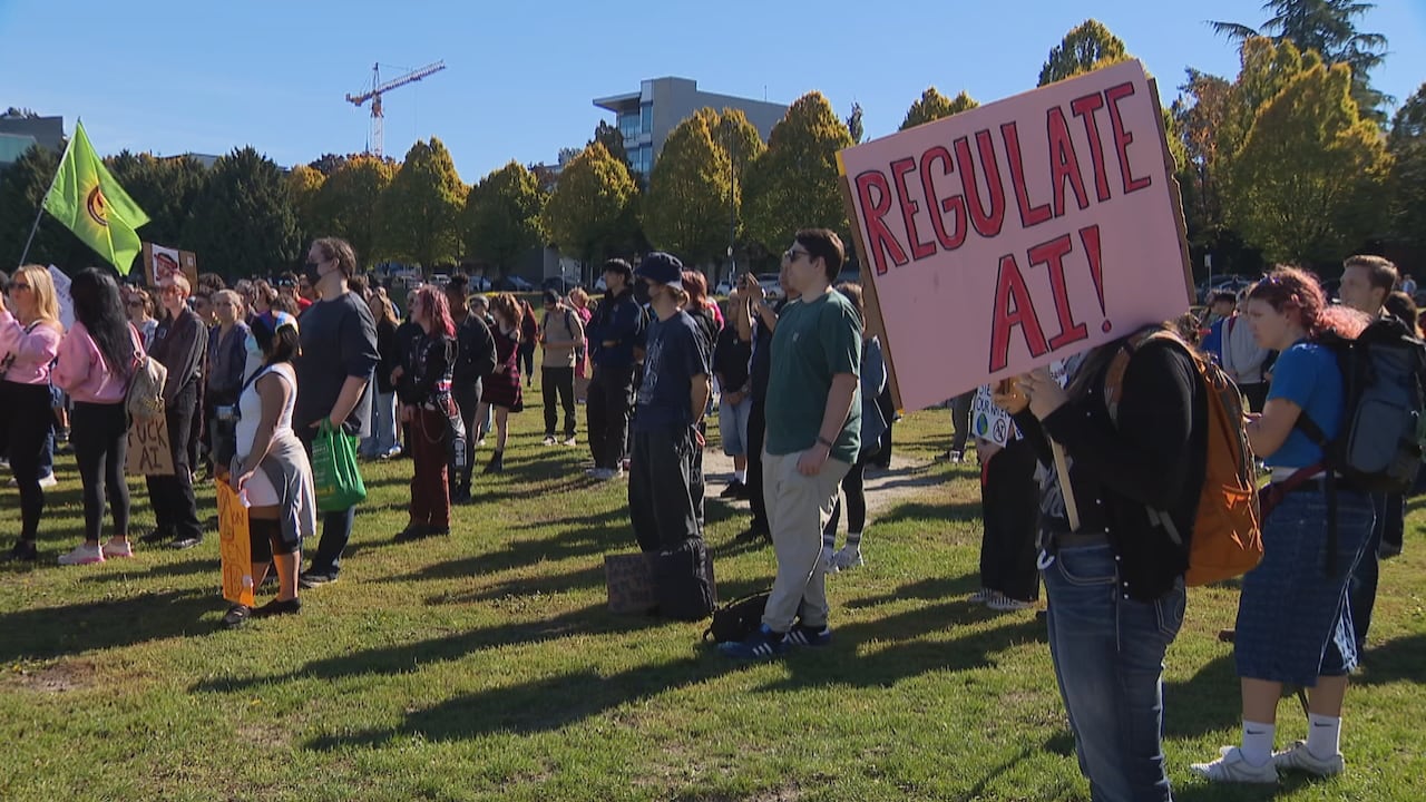 A crowd gathered on grass at an outdoor protest, one person holding a large pink placard reading 'Regulate AI!' with university buildings and a construction crane behind them.